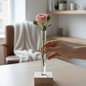 May include: A clear glass test tube vase set into a square light-coloured wooden base. A single pink rose with several green buds and a green stem is placed inside the glass tube, which is filled with water to hold the flower upright.