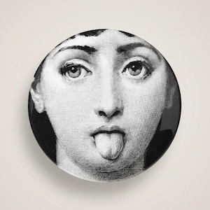 May include: A black and white decorative plate featuring a detailed illustration of a face with the tongue sticking out. The plate has a glossy finish and a round shape. The face is centered on the plate.