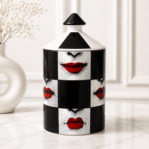 May include: A cylindrical ceramic canister with a black and white checkered pattern featuring repeating graphic prints of red lips. The container has a matching black and white lid with a pointed top, creating a bold, high-contrast visual design.