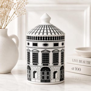 May include: A cylindrical white ceramic jar with a lid, featuring a black architectural line drawing of a building facade with windows, doors, and a tiled roof pattern. The jar has a pointed lid top and a smooth, glossy finish.