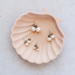 Picture shows 4 pairs of circular, white coloured pearl stud earrings, with silver post and scroll back/butterfly fixings. The earrings are of various sizes and sit on a shell shaped plate.
