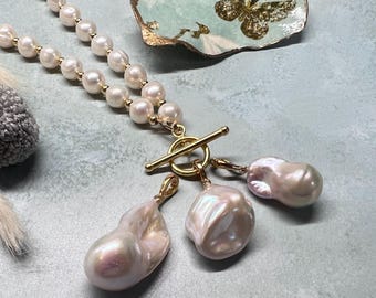 Könnte beinhalten: Eine Perlenkette mit goldenem Verschluss und drei großen, unregelmäßig geformten Perlenanhängern. Die Kette hat zwei Stränge aus kleinen, runden Perlen. Der goldene Verschluss ist im Stäbchen- und Ring-Stil gehalten. Die Perlen haben einen weichen, irisierenden Glanz.