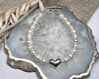 Könnte beinhalten: Ein weißes Perlenarmband mit einem silbernen Herz-Anhänger. Das Armband liegt auf einer weißen und goldenen Achat-Scheibe.