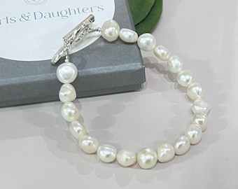 Könnte beinhalten: Ein weißes Perlenarmband mit einem silbernen Verschluss. Das Armband liegt auf einer grauen Schachtel mit dem Text "Girls & Daughters" darauf.