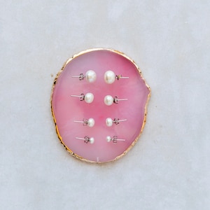 Picture shows 4 pairs of circular, white coloured pearl stud earrings, with silver post and scroll back/butterfly fixings. Earrings are of varying sizes. The earrings are elegantly placed on a pink shell.