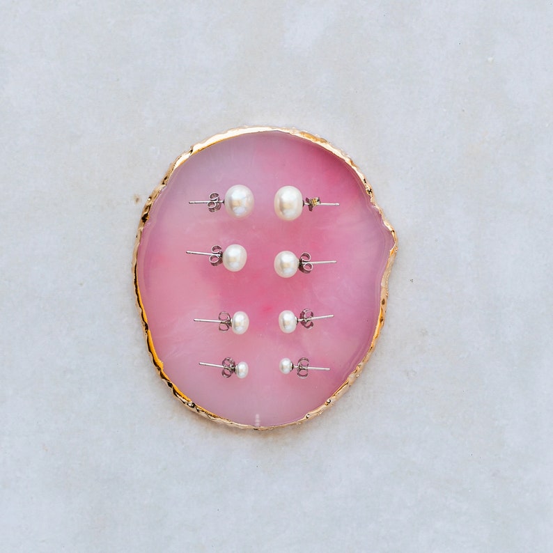 Picture shows 4 pairs of circular, white coloured pearl stud earrings, with silver post and scroll back/butterfly fixings. Earrings are of varying sizes. The earrings are elegantly placed on a pink shell.