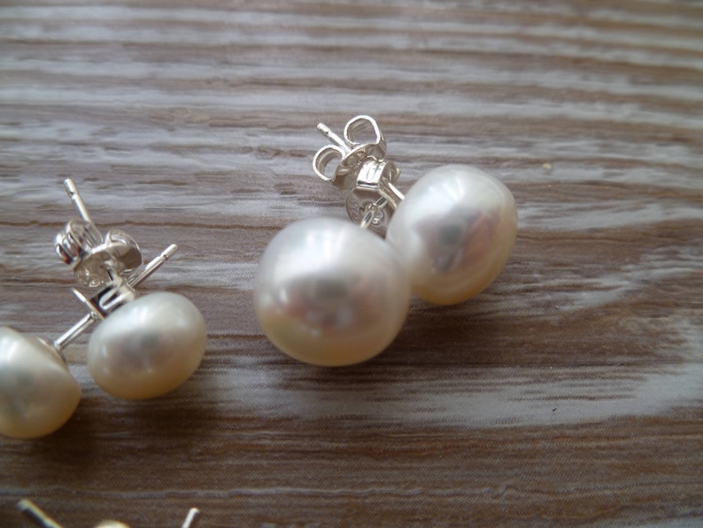 Picture shows a pair of circular, white coloured pearl stud earrings, with silver post and scroll back/butterfly fixing. A smaller pair of the same type of earring also feature in the picture.