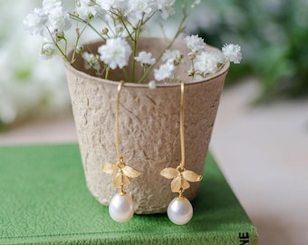 Könnte beinhalten: Goldene Ohrringe mit Perlen-Tropfen und einem zarten Schmetterlingsdesign. Die Ohrringe werden in einem braunen Papiertopf mit weißen Gipskrautblüten präsentiert.