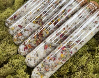 May include: Six clear glass tubes filled with white granules and dried flowers, including red and yellow petals, are arranged diagonally. The tubes are capped with corks and rest on a bed of green moss, creating a natural, spa-like aesthetic.