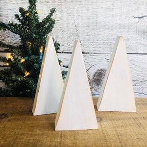 May include: Three white wooden triangle Christmas trees with a distressed white paint finish. The trees are arranged in a row on a wooden surface.