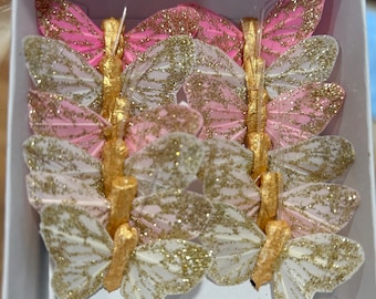 May include: A set of six glittery butterfly embellishments in pink and white with gold accents. The butterflies are made of feathers and have a delicate, feminine look.
