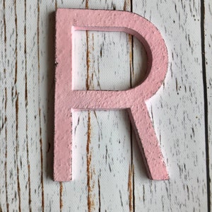 May include: A pink wooden letter R. The letter is painted a light pink color and has a slightly distressed look. The letter is sitting on a white wooden background.