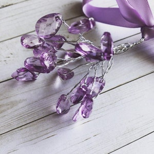 May include: A cluster of purple crystal beads dangle from a silver wire, attached to a purple ribbon. The beads are various shapes and sizes, and they are arranged in a cascading pattern.