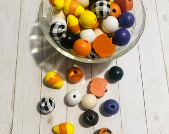 May include: A bowl of assorted wooden beads in orange, yellow, black, white, and purple. The beads are shaped like pumpkins, candy corn, and buffalo plaid.