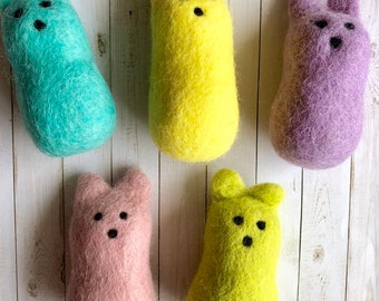 May include: Five felted wool animal figures in shades of teal, yellow, purple, pink, and chartreuse. Each has a rounded body, small ears, and three black dots for facial features. The figures are arranged on a light wood-grain surface.