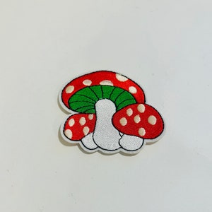 May include: Embroidered patch featuring three red and white mushrooms with white spots. The top mushroom has a green cap.