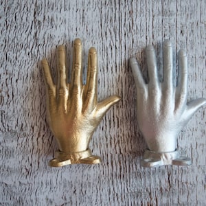 May include: Two decorative hand-shaped hooks, one gold and one silver, mounted on a white wooden surface.