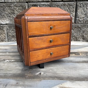 May include: A small wooden chest with three horizontal drawers, each featuring a single small metal knob. The unit has a beveled top and vertical slat detailing on the side, constructed from a medium-toned brown wood with a visible grain texture.