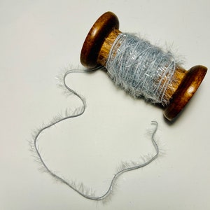 May include: A spool of silver tinsel yarn wound around a wooden spool. The yarn is fluffy and has a shimmery finish.