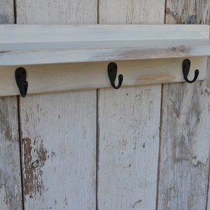 May include: White wooden shelf with three black metal hooks. The shelf has a distressed finish and is mounted on a white wood wall.