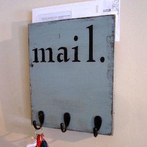 May include: A light blue wooden mail holder with the word "mail" in black lettering. The holder has four hooks on the bottom for hanging keys. There is a white envelope with a stamp in the top slot.