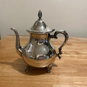 May include: A silver-colored teapot with a curved spout and ornate handle. The lid is topped with a decorative finial. The teapot sits on three small feet and reflects the surrounding environment.