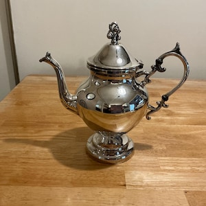 May include: A silver-colored teapot with a decorative handle and spout. The teapot has a bulbous body and sits on a pedestal base. The lid is topped with a decorative finial. The teapot is highly reflective, showing the surrounding environment.