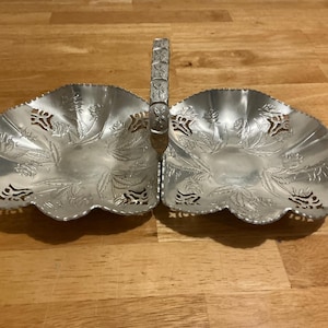 May include: A silver-colored, two-section serving dish with a handle. Each section has a scalloped edge and is decorated with floral patterns. The handle is also decorated with a repeating pattern. The dish is made of metal.