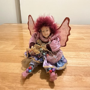 May include: A porcelain doll with maroon hair and pink wings, wearing a layered outfit in shades of pink, purple, and blue. The doll holds a gold-toned decorative string with small colorful beads and wears boots adorned with tiny bells.