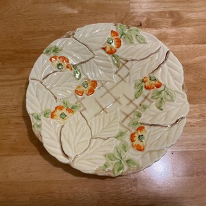 May include: A round ceramic plate featuring a raised, embossed pattern of cream-colored leaves and a central lattice design. The plate is decorated with several small, hand-painted orange flowers with green centers and light green foliage accents.