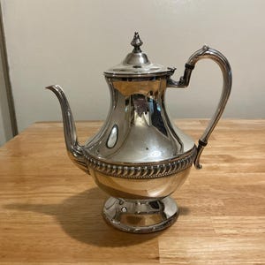 May include: A silver-colored teapot with a curved spout and handle. The teapot has a decorative beaded band around the middle and a lid with a small finial. It sits on a round base. The teapot is likely made of metal.