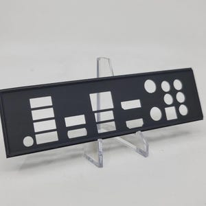 May include: A black computer I/O shield with various rectangular and circular cutouts. The shield is displayed on a clear acrylic stand against a white background. The cutouts are for ports and connectors.