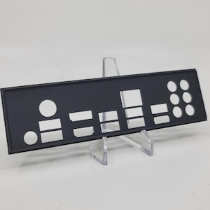 May include: A black computer I/O shield with various cutouts for ports and connectors. The rectangular plate is supported by a clear plastic stand, showcasing its design and features. The shield is designed for a computer's rear panel.