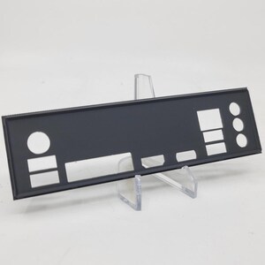 Puede incluir: Un panel trasero de computadora negro con varias formas recortadas para puertos y conectores. El panel rectangular tiene un acabado mate y se muestra sobre un soporte de acrílico transparente sobre un fondo blanco. Tiene aberturas rectangulares y circulares.
