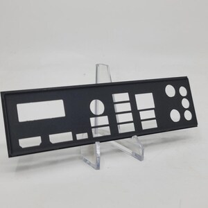 May include: A black computer component with various cut-out shapes and openings. The rectangular piece is supported by a clear plastic stand, and is designed to fit the back panel of a computer case. The component has several rectangular and circular openings.