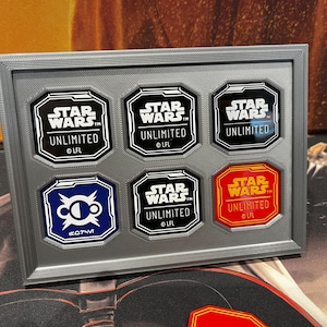 Può includere: Un espositore incorniciato con sei pedine di gioco ottagonali di Star Wars Unlimited. I pezzi sono neri, bianchi, blu e rossi, con il logo Star Wars Unlimited. La cornice è grigia.