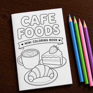 May include: A white mini coloring book titled CAFE FOODS with black line art illustrations of a coffee cup, a slice of cake with a strawberry, and a croissant on plates. Four colored pencils in blue, green, pink, and purple lie beside the book.