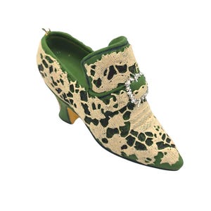 May include: A decorative shoe ornament in a vintage style. The shoe is green with a cream-colored lace overlay. A sparkling rhinestone buckle adorns the top. The heel is yellow and the shoe has a pointed toe.