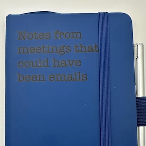 Puede incluir: Se muestra un cuaderno azul con el texto "Notes from meetings that could have been emails". Un bolígrafo plateado se sujeta con una banda elástica azul. El cuaderno está cerrado y tiene una superficie lisa.