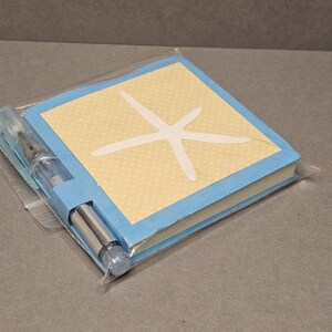 Op de afbeelding: Een klein, vierkant notitieboekje met een lichtblauwe kaft en een lichtgele middenkant met een wit zeesterontwerp. Een doorzichtige pen is aan de zijkant van het notitieboekje bevestigd. Het notitieboekje is verpakt in een doorzichtige plastic zak.
