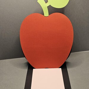 May include: A large, red apple cutout with a green leaf and stem. The apple is positioned above a black rectangle, which holds a square of pink paper. The background is a neutral gray.