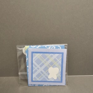 May include: A clear plastic bag containing a blue and white card with a white butterfly design. The card has a blue border and a plaid pattern in the center. The background is a solid gray.