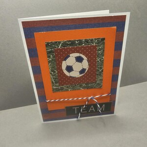 Pode incluir: Um cartão de felicitações feito à mão com um tema de futebol. O cartão apresenta um desenho de bola de futebol no centro, rodeado por camadas de papel laranja, castanho e azul. A palavra "TEAM" é exibida na parte inferior, e o cartão é amarrado com uma corda branca e azul.