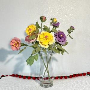 May include: A clear glass vase holds a bouquet of handmade flowers. The flowers are in shades of yellow, pink, and purple, with green leaves. The vase sits on a white surface with a red pom-pom trim.