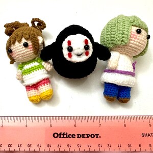 以下が含まれることがあります： 手作りの3つの編み人形。1つの人形は茶色の髪で、緑と白のストライプのシャツ、ピンクのショートパンツ、黄色の靴を履いています。別の人形は緑色の髪、白いトップス、青いパンツ、茶色の靴を履いています。白い顔の黒い人形が中央にあります。
