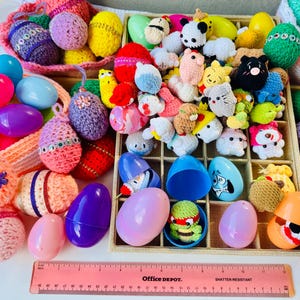 May include: A collection of colorful Easter eggs and decorative items. The image features plastic eggs, crocheted eggs, and small stuffed animal figures in a variety of colors. A pink Office Depot ruler is at the bottom.