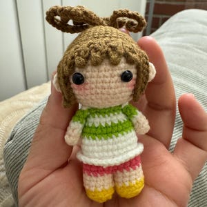 以下が含まれることがあります： 手作りのかぎ針編み人形。茶色の髪を2つの団子ヘアにしています。人形は、緑と白のストライプのトップス、ピンクのショートパンツ、黄色の靴を履いています。人形は黒い目とピンクの頬をしています。