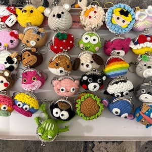 May include: A collection of colorful, crocheted coin purses in various animal and character designs. The purses feature metal clasps and key rings. Designs include a bear, cat, dog, frog, and other whimsical creatures. The purses are displayed in a white box.