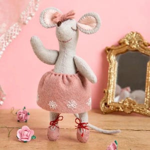 May include: A light gray felt mouse figurine with a pink bow on its head, wearing a dusty pink skirt with white embroidered flower details and red ribbon ballet slippers. The mouse has closed eyes and a long tail, standing upright.