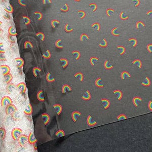 May include: A roll of white fabric with a rainbow pattern next to a sheet of black fabric with the same rainbow pattern. The rainbows are in red, orange, yellow, green, blue, and purple.